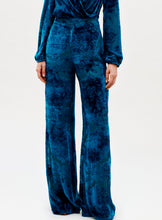 Load image into Gallery viewer, A person wears the Como Pant—high-waisted, wide-leg velvet pants in deep blue with a subtle textured pattern—paired with a matching long-sleeve top, set against a plain, light background.