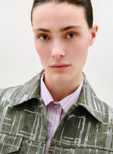 Load image into Gallery viewer, A person with light skin, straight brown hair slicked back, and hazel eyes looks at the camera. They wear the Lesath Jacket—a green plaid piece with a shiny finish—over a pink and white striped collared shirt. The background is plain and light.