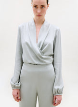 Load image into Gallery viewer, A person with a neutral expression stands against a plain light background, wearing the light gray, long-sleeved Tea Blouse with a deep V-neck and matching high-waisted pants. Their arms are relaxed by their sides.