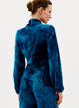 Load image into Gallery viewer, A woman with light skin and brown hair in a bun is seen from the back, wearing the Tea Blouse in deep blue Italian velvet with long sleeves, standing against a plain white background.
