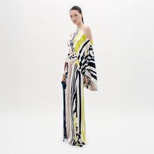 Load image into Gallery viewer, A woman stands in a white space wearing the Dotty Dress—a one-shoulder, floor-length gown with bold black, yellow, and red stripes—while holding a tasseled accessory.