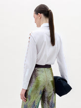 Load image into Gallery viewer, A woman stands against a plain background wearing the Idris Blouse with cut-out sleeves, paired with iridescent wide-leg pants, a large belt, and holding a folded black garment.