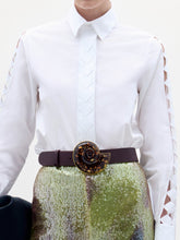 Load image into Gallery viewer, A woman stands against a plain background wearing the Idris Blouse with cut-out sleeves, paired with iridescent wide-leg pants, a large belt, and holding a folded black garment.