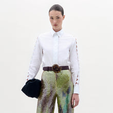 Load image into Gallery viewer, A woman stands against a plain background wearing the Idris Blouse with cut-out sleeves, paired with iridescent wide-leg pants, a large belt, and holding a folded black garment.