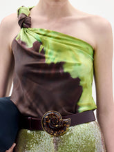 Load image into Gallery viewer, A woman stands against a plain background wearing the Yanet Blouse, a one-shoulder green and brown top, paired with sequined pants, a wide belt with a large buckle, and holding a black clutch.