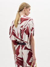 Load image into Gallery viewer, A woman poses against a plain white background wearing the Susanne Blouse, featuring a red and white abstract pattern, matched with coordinating pants and accessorized with a black belt with a gold buckle.