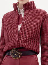 Load image into Gallery viewer, A woman stands against a plain background wearing the Zalta Jacket, a textured red jacket paired with a striped shirt, red pants, and a large black belt featuring an ornate buckle.