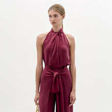 Load image into Gallery viewer, A woman stands against a plain background wearing the Shela Blouse, a sleeveless, high-neck magenta top with a tie detail at the waist, paired with a small handbag.