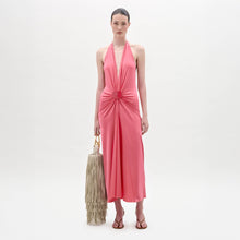 Load image into Gallery viewer, A woman stands against a plain background wearing the sleeveless pink Veracruz Dress with a gathered waist, holding a large beige fringed bag and wearing brown sandals.