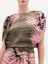 Load image into Gallery viewer, A woman stands against a plain background wearing the Alisa Blouse, featuring a pink and olive green pattern crafted from Italian fabric. She holds a brown bag and wears gold bracelets.