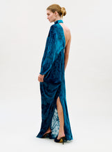 Load image into Gallery viewer, A woman in a low bun showcases the Anya Dress, a long blue velvet gown with an open back and high slit, paired with black heels against a plain light background.