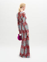 Load image into Gallery viewer, A person stands against a plain background wearing the Barisa Dress, a long, patterned red and blue fitted dress, and holding a small purple handbag.