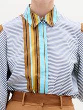 Load image into Gallery viewer, A person stands against a plain background wearing the oversized fit Belkis Blouse with balloon sleeves and stripes, paired with tan high-waisted pants, and holding a tan accessory in their left hand.
