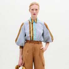 Load image into Gallery viewer, A person stands against a plain background wearing the oversized fit Belkis Blouse with balloon sleeves and stripes, paired with tan high-waisted pants, and holding a tan accessory in their left hand.