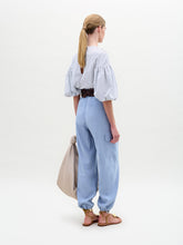 Load image into Gallery viewer, A woman stands against a plain background wearing a striped blouse with blue floral appliqué, the Darnell Pant in light blue, a brown belt with a large buckle, gold sandals, and holding a beige bag.