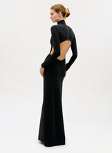 Load image into Gallery viewer, A person stands against a plain white background wearing the Birdy Dress, a long, fitted black gown featuring long sleeves and an open back, with their hair styled in a neat bun.
