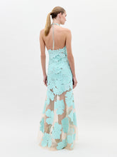 Load image into Gallery viewer, A woman wearing the Mab Dress Turquoise, a light blue floral-patterned halter neck dress with a sheer skirt, stands against a plain background, eagerly awaiting the pre-order ship-by date of February 1st.
