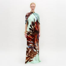 Load image into Gallery viewer, A woman stands against a white background wearing the Carlina Dress, a floor-length light blue maxi made from Italian fabric with a brown and red feather-like pattern and a matching draped shoulder cape.
