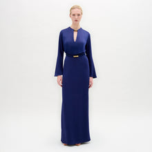 Load image into Gallery viewer, A person stands facing forward in the Cassy Dress—a long-sleeved, floor-length dark blue maxi with a keyhole neckline and black belt, crafted from luxurious Italian fabric—set against a plain white background.