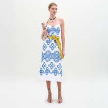 Load image into Gallery viewer, A woman stands against a plain background wearing the Ceres Dress, an embroidered linen midi with blue patterns and a yellow belt, crafted from fine Italian fabric. She pairs it with tan sandals and carries a beige shoulder bag.