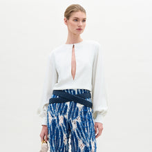 Load image into Gallery viewer, A woman poses against a plain background wearing the Covilha Blouse, featuring a keyhole neckline and balloon sleeves, styled with high-waisted blue patterned pants and holding a small beige bag.