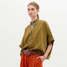 Load image into Gallery viewer, A woman stands sideways against a plain white background, wearing the Daia Blouse in olive green with a V-cut neckline, paired with loose orange pants and holding a textured yellow bag.
