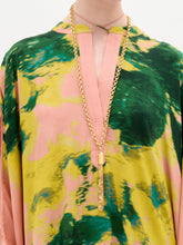 Load image into Gallery viewer, A woman stands against a white background wearing the Elea Tunic, an oversized 100% silk maxi dress with pink, yellow, and green patterns, paired with a beige bag and strappy heels.