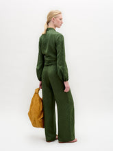 Load image into Gallery viewer, Person standing against a plain background wearing the green Elvie Blouse, a long-sleeved wrap top with wrap-around ties, matching pants, accessorized with a gold necklace and holding a gold handbag.
