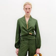 Load image into Gallery viewer, Person standing against a plain background wearing the green Elvie Blouse, a long-sleeved wrap top with wrap-around ties, matching pants, accessorized with a gold necklace and holding a gold handbag.