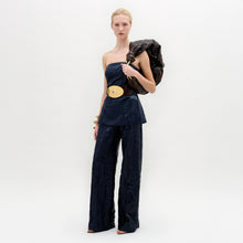 Load image into Gallery viewer, A woman stands against a white background wearing the Emine Pant—a high-waisted, wide-leg silhouette—paired with a dark strapless top, gold belt, bangles, and a textured bag slung over her shoulder.