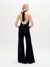 Load image into Gallery viewer, A person stands against a white background in the Enara Jumpsuit, a black halter-neck piece made from luxurious Italian fabric with wide-leg pants, accessorized with elegant gold bangles.