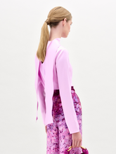 Load image into Gallery viewer, A woman poses against a plain background in the Gaby Blouse, a light pink silk long-sleeve top with a front slit, paired with high-waisted pink and purple patterned pants, a wide belt, and a small matching handbag.