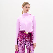 Load image into Gallery viewer, A woman poses against a plain background in the Gaby Blouse, a light pink silk long-sleeve top with a front slit, paired with high-waisted pink and purple patterned pants, a wide belt, and a small matching handbag.