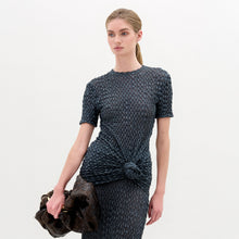 Load image into Gallery viewer, A woman with blonde hair in a low ponytail wears the Gen Tshirt, a textured, fitted, dark short-sleeve top with knot detail, and holds a large dark ruched bag against a plain white background.