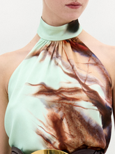 Load image into Gallery viewer, A woman stands against a plain white background wearing the Geovanna Blouse—a sleeveless, high-neck halter top featuring a brown and light blue abstract pattern—paired with a wide belt and matching skirt, made in Colombia.