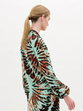 Load image into Gallery viewer, A woman stands against a plain white background wearing the Honey Blouse, a long-sleeved top featuring green, black, and orange abstract patterns with a chic front twist detail.
