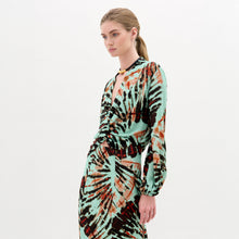 Load image into Gallery viewer, A woman stands against a plain white background wearing the Honey Blouse, a long-sleeved top featuring green, black, and orange abstract patterns with a chic front twist detail.
