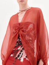 Load image into Gallery viewer, A woman stands against a plain background wearing the Idra Blouse, a sheer, long-sleeved top with a deep V neckline, layered over a patterned shirt and paired with a matching red skirt.