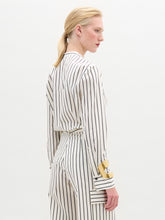 Load image into Gallery viewer, A person stands against a plain background wearing the Isabell Blouse, a white long-sleeve top with black lace and delicate embroidery down the front, paired with pants featuring black vertical stripes.