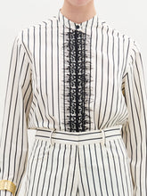 Load image into Gallery viewer, A person stands against a plain background wearing the Isabell Blouse, a white long-sleeve top with black lace and delicate embroidery down the front, paired with pants featuring black vertical stripes.