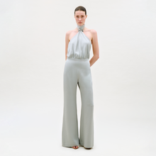 Load image into Gallery viewer, A woman stands against a plain light background wearing the Imperio Jumpsuit—a light gray, sleeveless halter-neck design with wide-leg pants. Her hair is pulled back and her hands are behind her back.