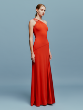 Load image into Gallery viewer, A woman in a Kanya Dress Rouge, featuring a red, one-shoulder design and floor-length cut, stands against a plain blue background. Her hair is pulled back as she calmly stands with her arms by her sides, appearing to wait for an important date such as August 19th or September 2nd.