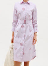 Load image into Gallery viewer, A person wears the Kendi Dress—a belted, knee-length, button-up dress with purple stripes, floral prints, and long sleeves—while holding a white textured bag against a plain white background.