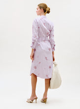 Load image into Gallery viewer, A woman stands with her back turned, wearing the Kendi Dress—a light pink striped shirt dress with floral prints. She carries a large white textured bag and wears beige high-heeled sandals. Her hair is styled in a neat bun against a plain light background.