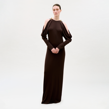 Load image into Gallery viewer, A woman stands against a plain light background, wearing the Kilian Dress—a long, dark brown dress featuring cold shoulder cutouts and gold-accented shoulders. Her hair is pulled back and her hands are clasped in front of her.