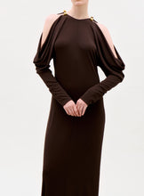 Load image into Gallery viewer, A person wearing the Kilian Dress, a long dark brown gown with draped cold-shoulder sleeves and gold clasp details at the shoulders, stands against a plain light background.