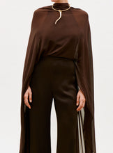 Load image into Gallery viewer, A person wears the Ligia Jumpsuit, a dark brown, semi-sheer piece with wide-leg pants, styled with a gold snake-shaped choker necklace. The background is plain white.