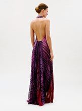 Load image into Gallery viewer, A woman stands facing away, wearing the Lucia Dress—a full-length, backless halter dress in purple and red abstract patterns. Her hair is styled in a neat bun against a plain white background.