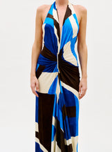 Load image into Gallery viewer, A woman wears the Malek Dress—a sleeveless, halter-neck style with a deep V-neckline, twist detail at the waist, and a bold blue, black, and beige geometric pattern—set against a plain white background.