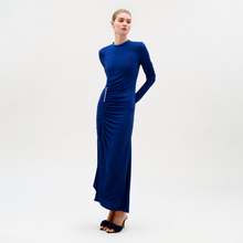 Load image into Gallery viewer, A woman stands against a plain background wearing the floor-length, long-sleeved Mara Dress in royal blue with ruched waist and center details, paired with black open-toe heels. Her hair is pulled back, and her hands are behind her back.